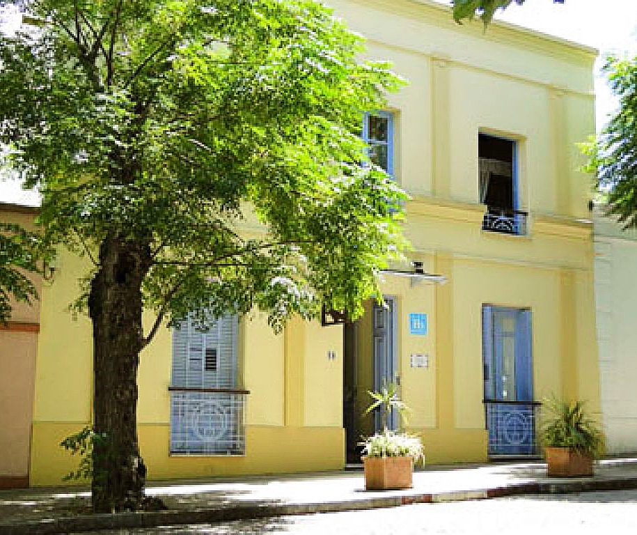 Fachada amarilla de la Posada del Ángel con persianas celestes y un árbol frente a la puerta